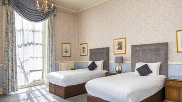 A hotel room with two beds, a large window with curtains, a chandelier, and framed pictures on the walls.