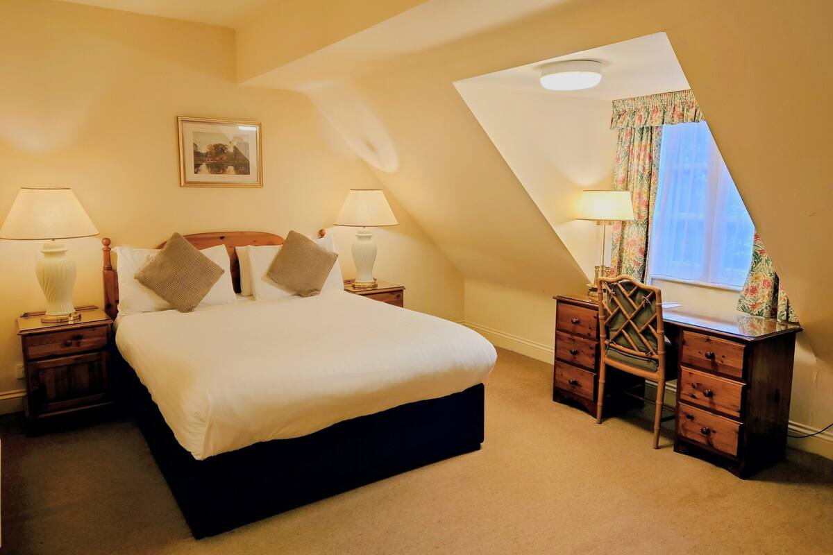 A bedroom with a large bed, two bedside tables with lamps, a dresser, a framed picture on the wall, and a window with curtains.