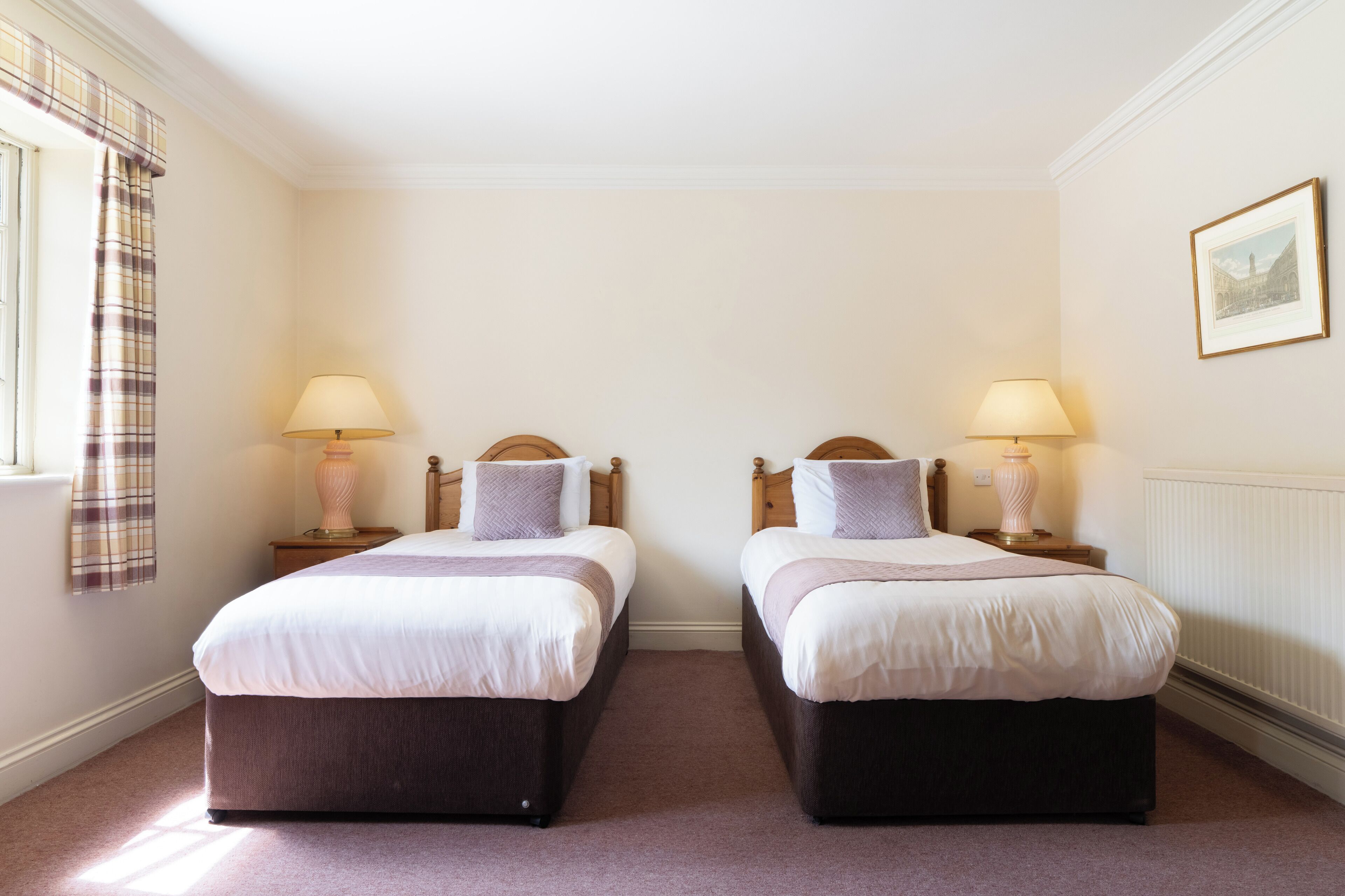 Una habitación con dos camas, cada una con una mesita de noche y una lámpara. La habitación tiene una ventana con cortinas y un cuadro en la pared.