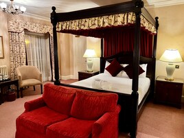 Una cama con dosel con ropa de cama roja y blanca, dos mesitas de noche con lámparas, un sillón tapizado en rojo y una silla con estampado floral.