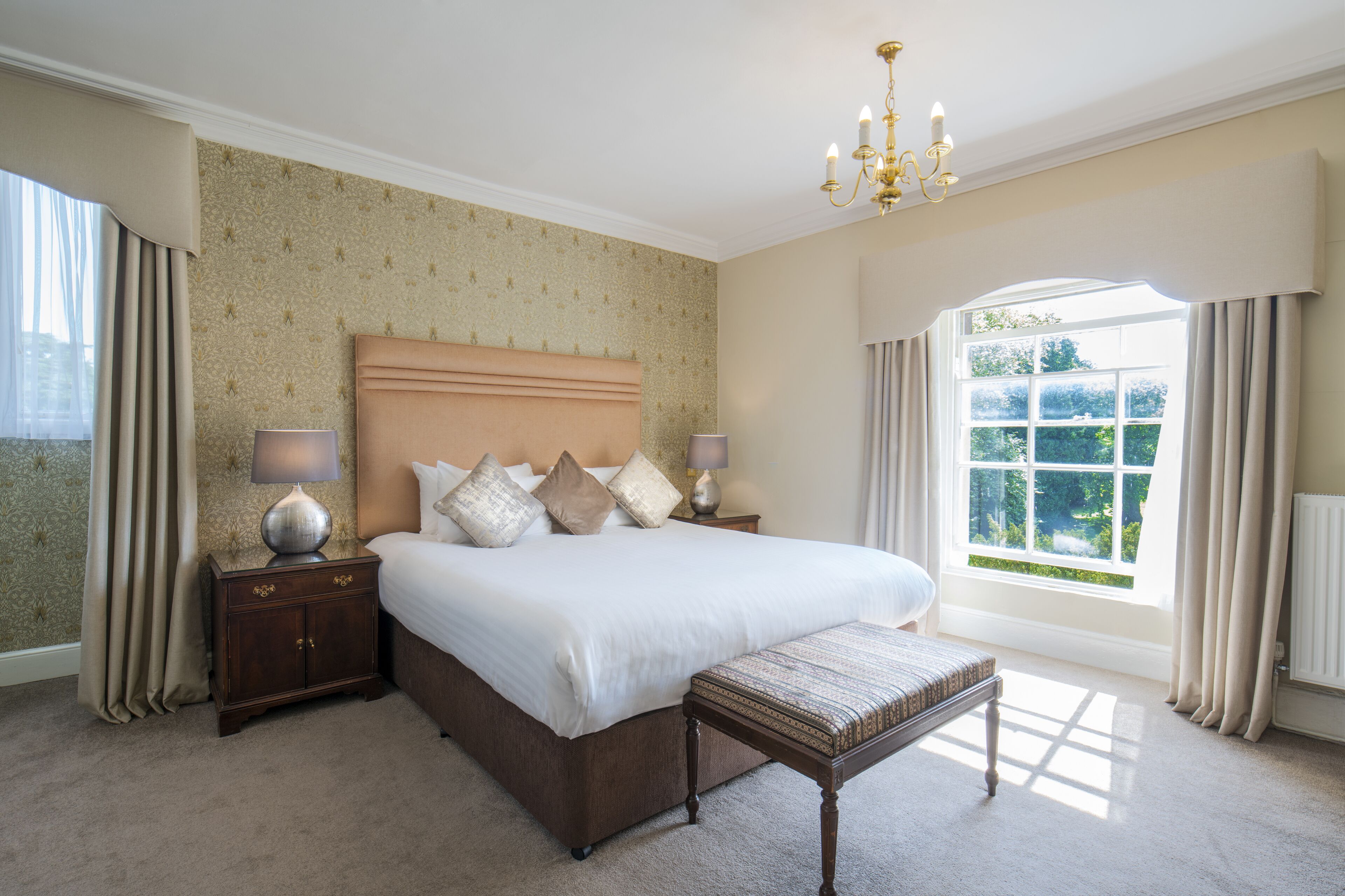 A bedroom with a large bed, bedside tables, a bench, a chandelier, and a window with curtains.