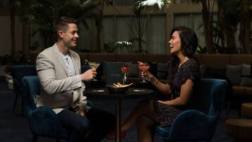 Um casal sentado à mesa com uma bebida e um vaso de flores, em um quarto com um sofá e plantas ao fundo.