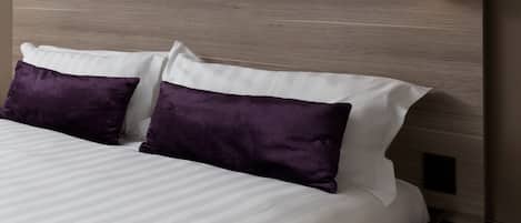 A neatly made bed with white and purple pillows, a wooden headboard, and two wall-mounted light fixtures.