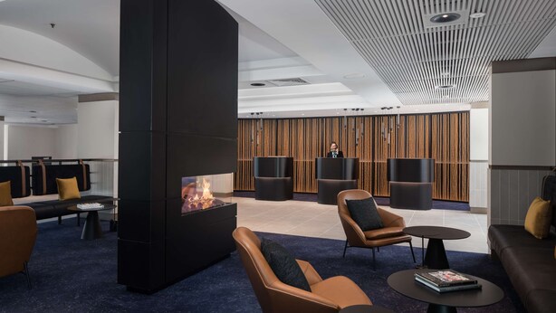 A modern lounge area with leather sofas, a central fire feature, and a wooden panel wall.