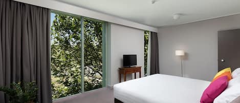 Une chambre d’hôtel moderne avec un grand lit, un téléviseur posé sur une table de chevet en bois, une plante en pot et une vue sur la verdure grâce à de grandes fenêtres du sol au plafond.