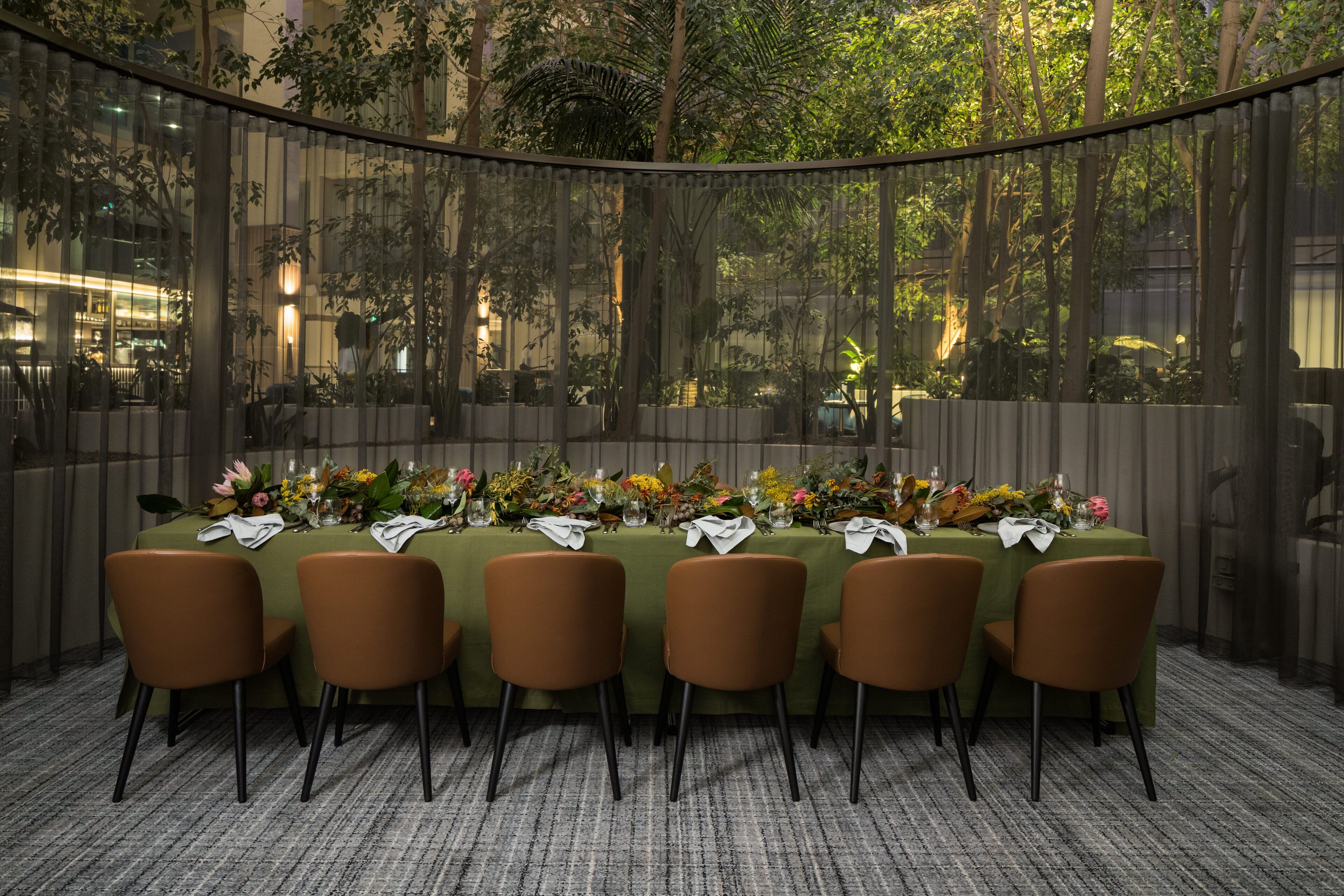 Comedor con una larga mesa dispuesta para seis personas, rodeada de sillas, con vista a árboles y edificios a través de un panel de vidrio.