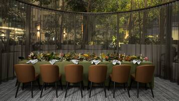 A dining area with a long table set for six, surrounded by chairs, with a view of trees and buildings through a glass partition.