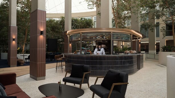 A modern bar area with a curved counter, seating with black and wooden chairs, and a high ceiling with natural light.