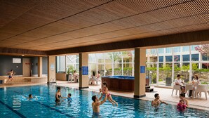 An indoor swimming pool with clear water, surrounded by a covered area with seating and a view of the outdoors. - Rydges Canberra (Forrest)