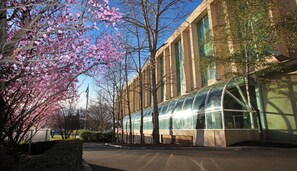 A building with a modern glass structure, surrounded by trees with pink blossoms, and a clear blue sky.