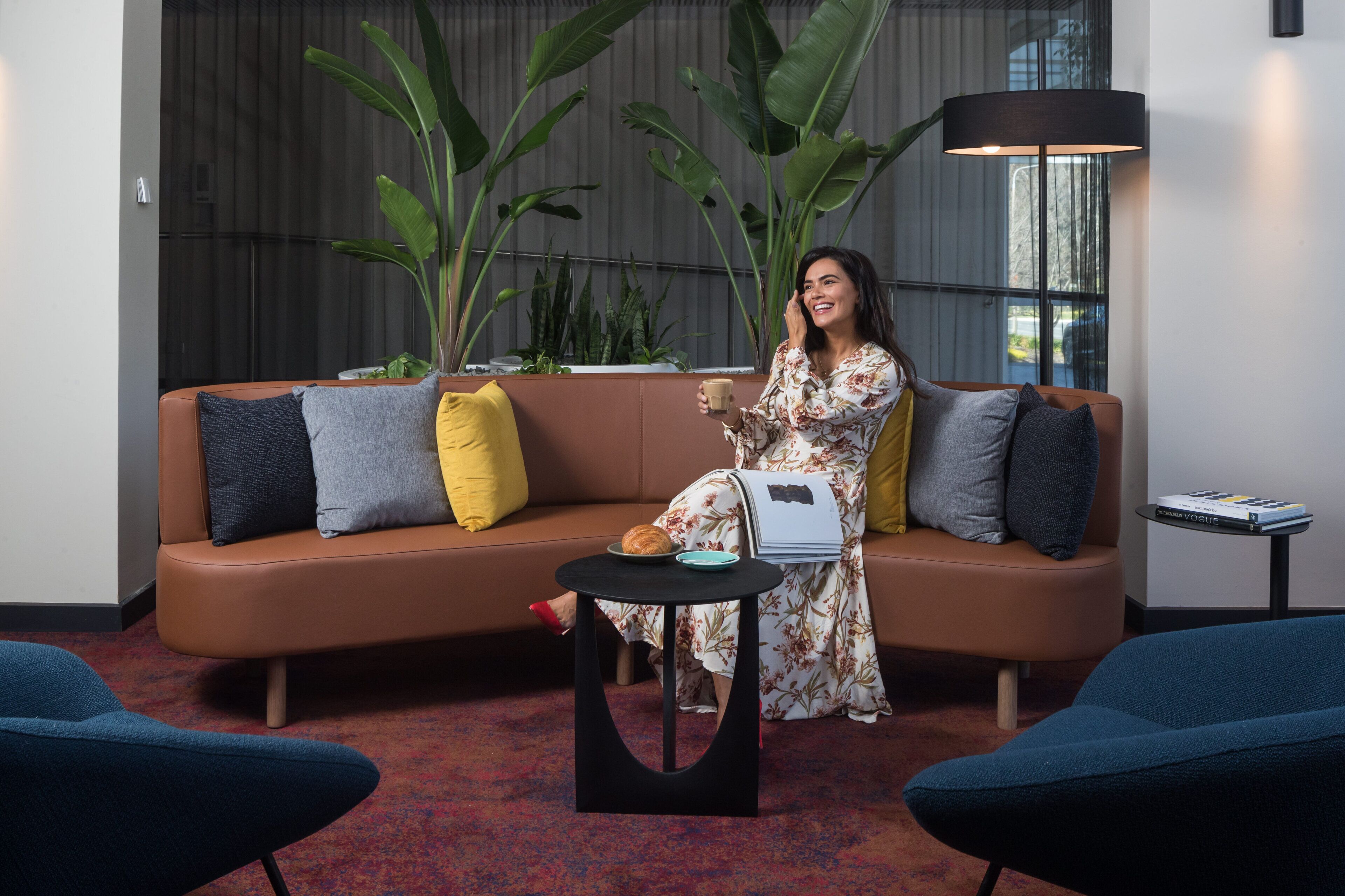 A woman sitting on a brown sofa with a coffee cup, surrounded by large green plants, a black lamp, and a small table with a book.