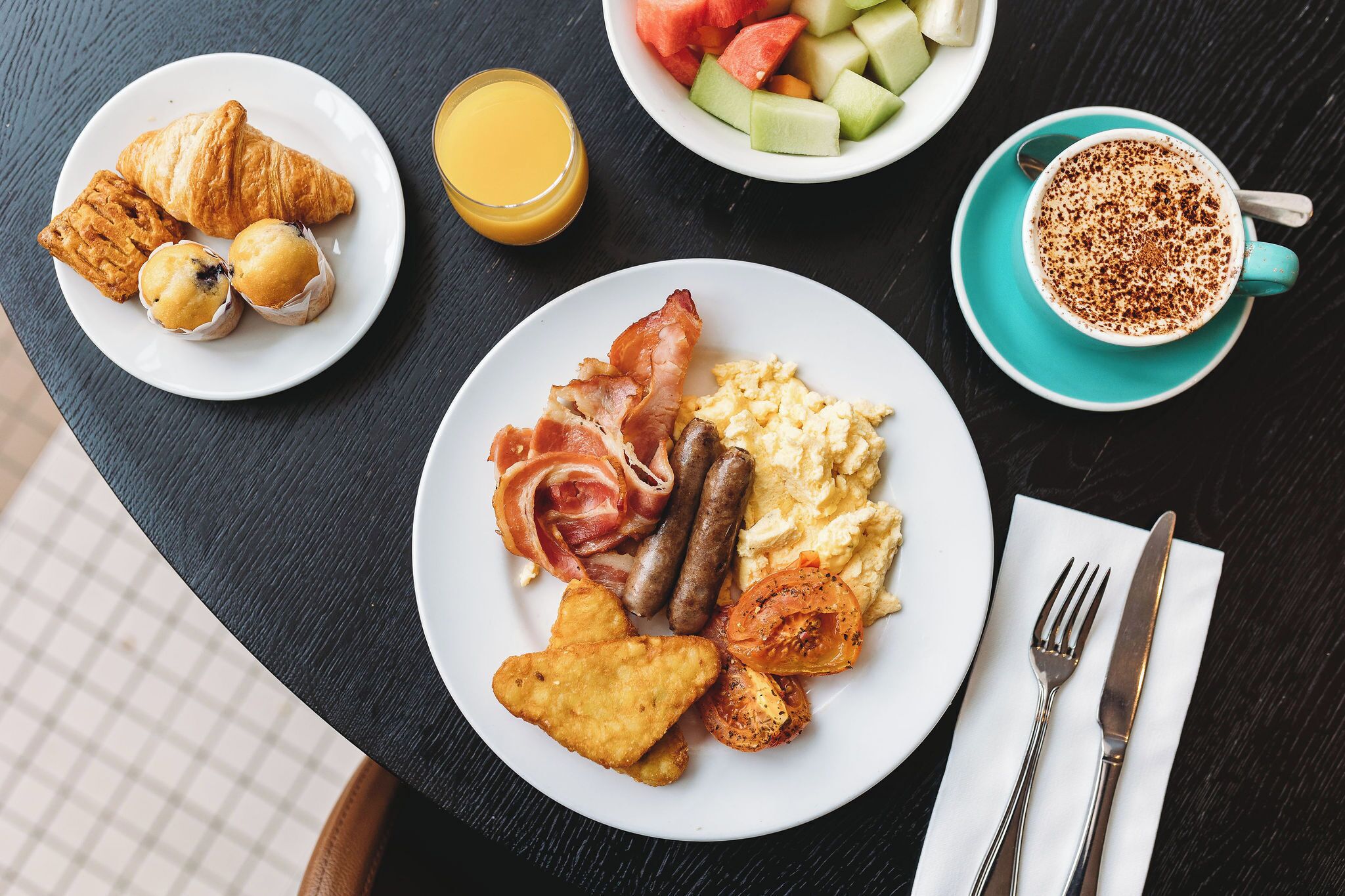 A breakfast spread on a table with croissants, muffins, bacon, sausages, scrambled eggs, toast, and a bowl of fruit.