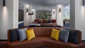 A modern lobby with a curved brown sofa, two yellow and two gray pillows, a black coffee table, and a seating area with a sofa and chairs. - Rydges Canberra (Forrest)