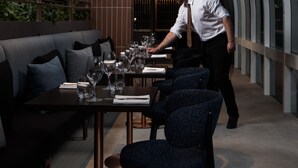 A modern restaurant with a high ceiling and curved glass walls. There are tables set with wine glasses and plates, and a waiter is attending to a table.