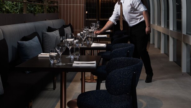 A modern restaurant with a high ceiling and curved glass walls. There are tables set with wine glasses and plates, and a waiter is attending to a table. - Rydges Canberra (Forrest)