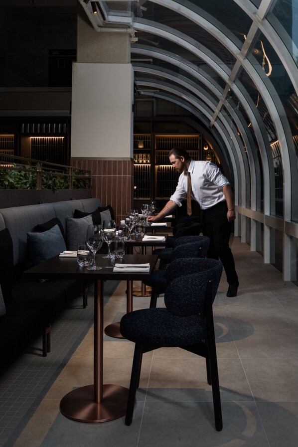 Un restaurant moderne avec un plafond haut et des murs en verre incurvés. On y trouve des tables dressées avec des verres à vin et des assiettes, et un serveur s’occupe d’une table.
