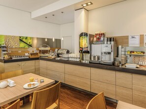 A modern breakfast area with a self-service counter, coffee machines, and a seating area with a table and chairs. - ibis Bamberg Altstadt (Bamberg)