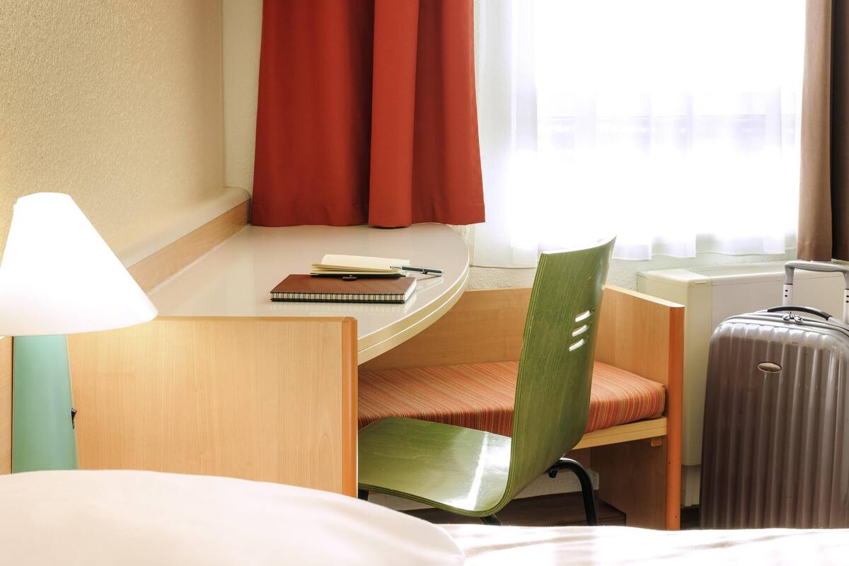 A hotel room with a bed, a desk with a lamp, a chair, a notebook, a pen, a suitcase, and a window with curtains.
