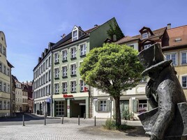 A cobblestone street with a statue, a green building with an 'ibis' hotel sign, and traditional European architecture.
