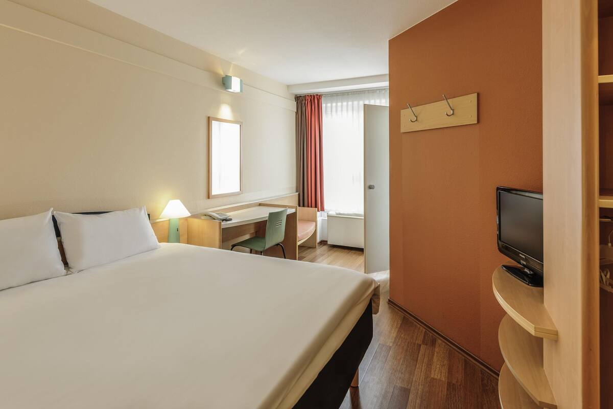 A hotel room with a large bed, a desk, a chair, a television, and a window with curtains.