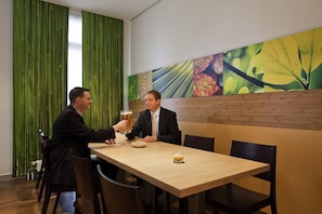 Two people seated at a wooden table in a restaurant with a green curtain backdrop and a large botanical wall mural. - ibis Bamberg Altstadt (Bamberg)