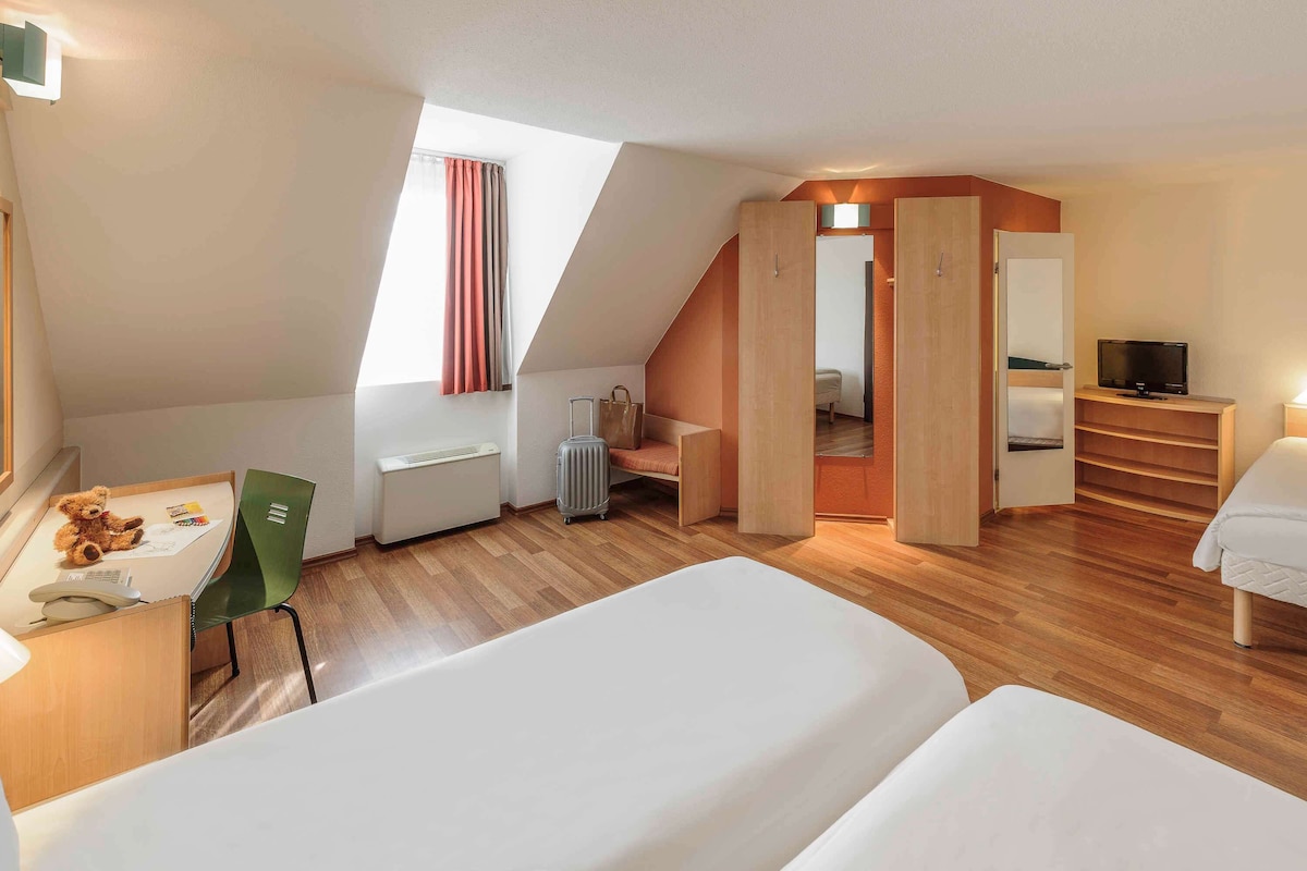 A modern, well-lit room with a sloped ceiling, a desk with a chair, a bed, a TV, and a wardrobe.