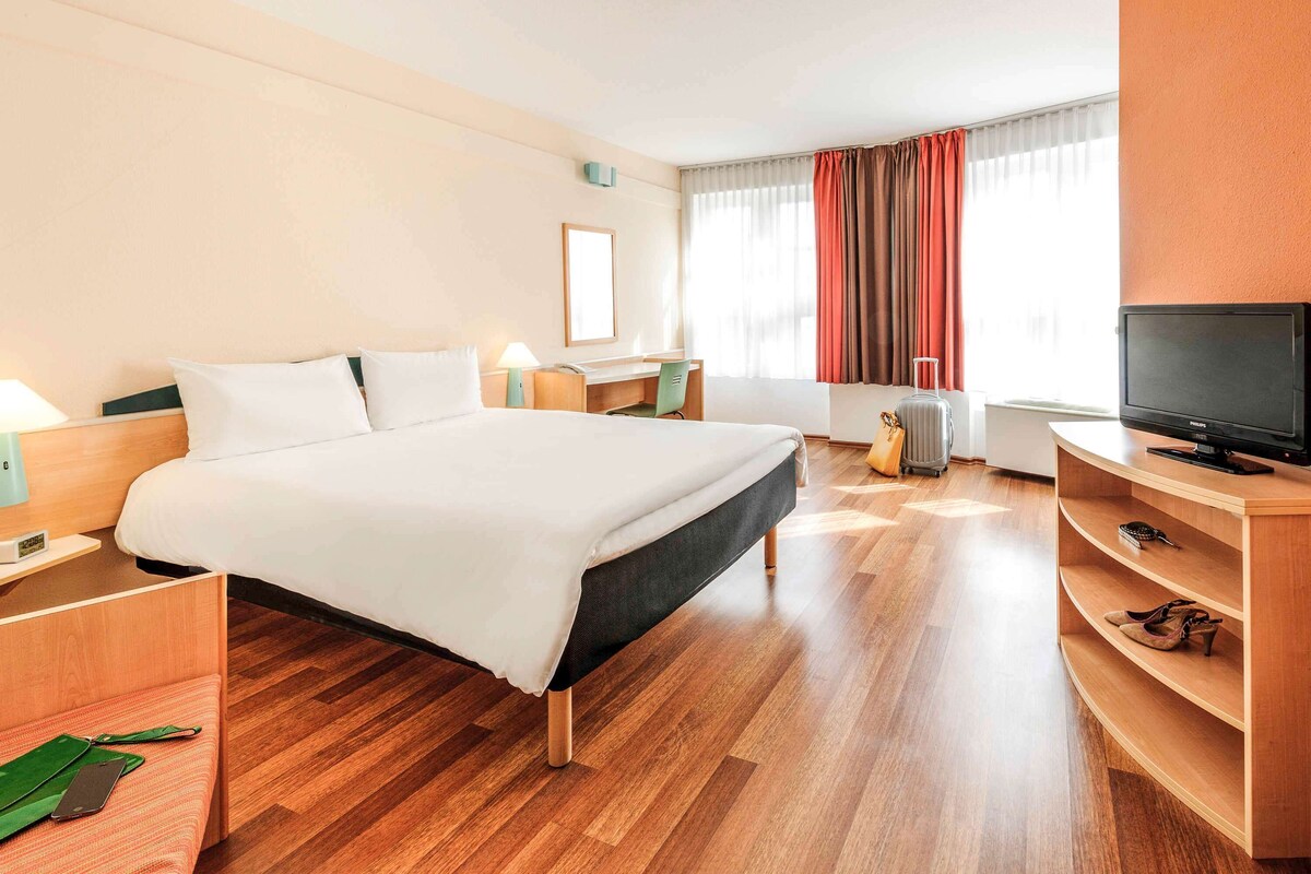 A hotel room with a large bed, a desk, a television, and a suitcase.