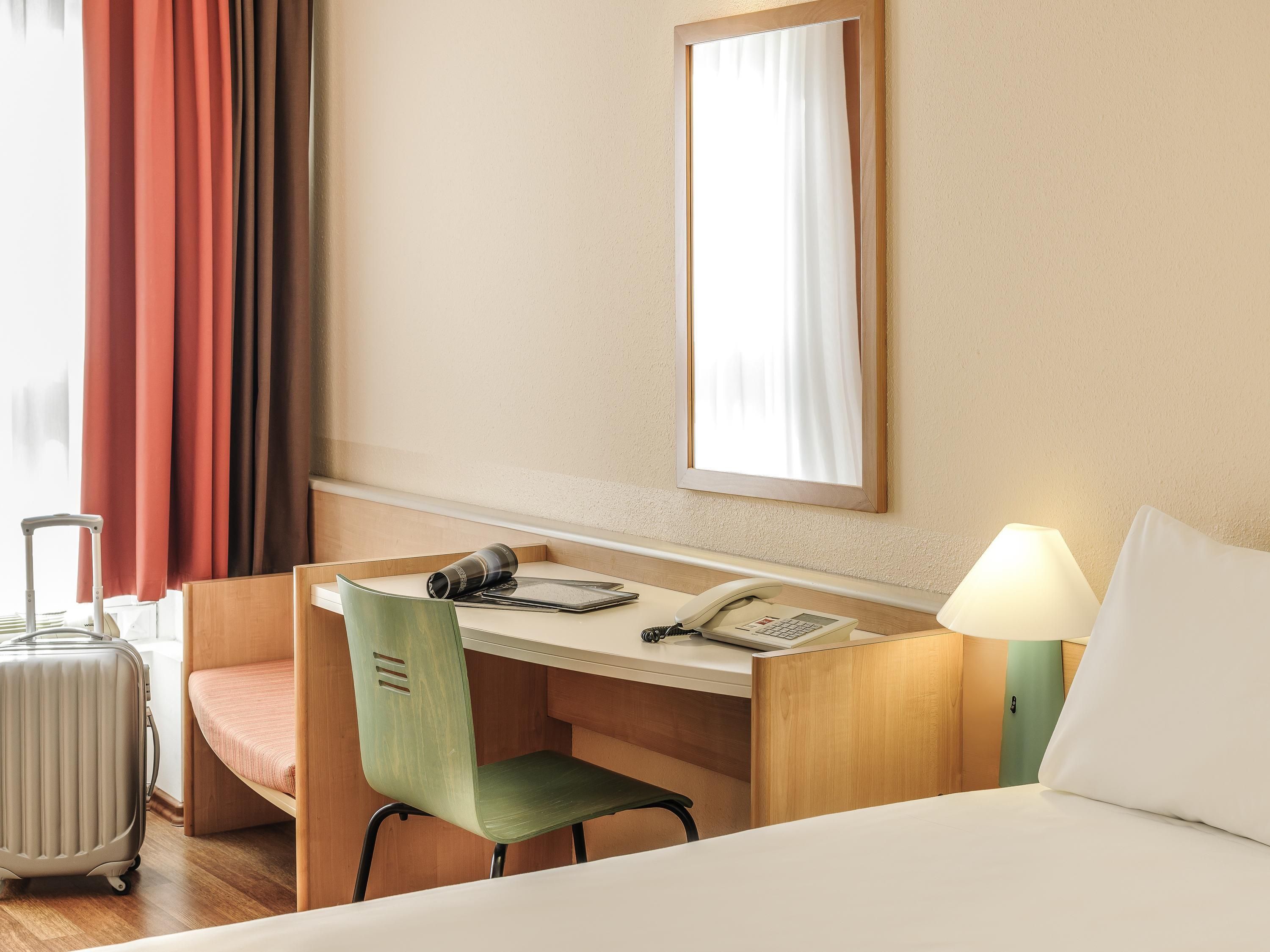 Habitación de hotel con una cama, un escritorio con silla, una lámpara, un teléfono y una maleta.