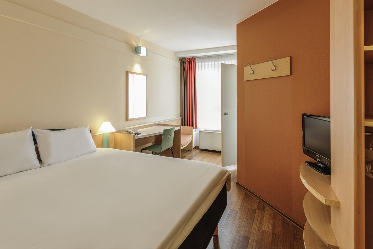 A hotel room with a large bed, a desk, a chair, a television, and a window with curtains.
