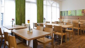 A modern dining area with wooden tables and chairs, large windows with green curtains, and a wall with a colorful abstract painting.