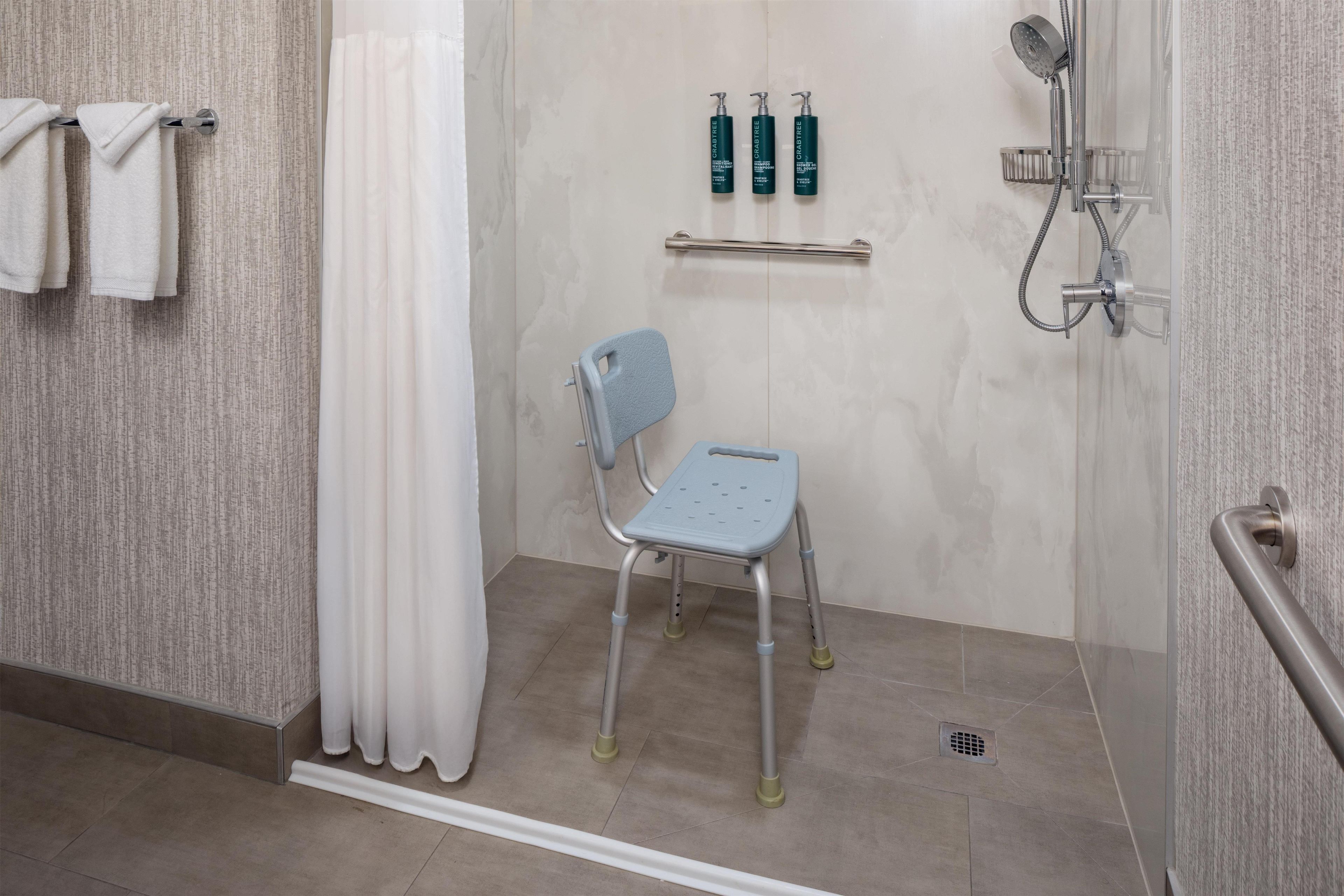 A bathroom with a shower, a grab bar, a wheelchair-accessible chair, and toiletries on a shelf.