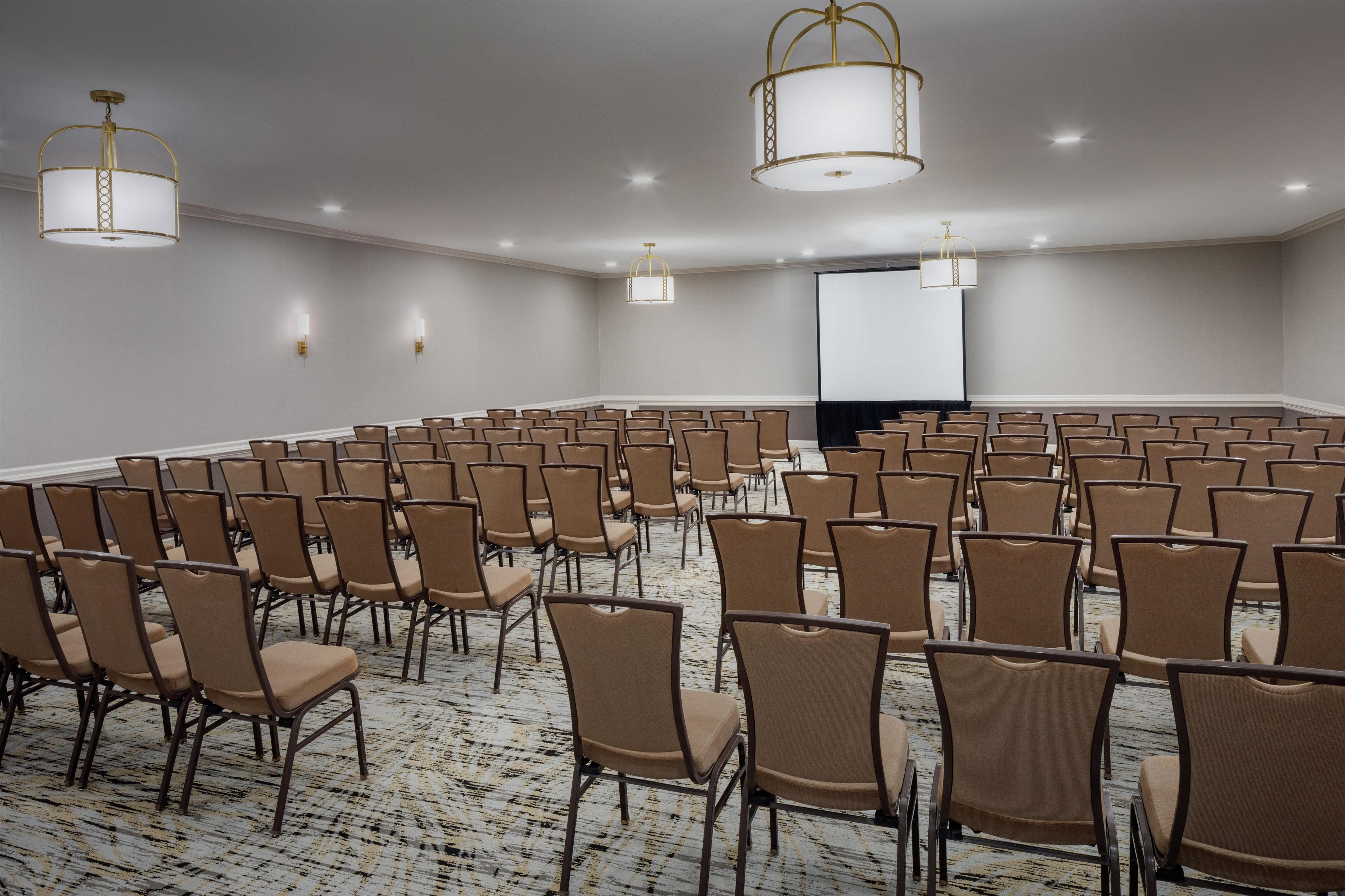 Un salón de conferencias con hileras de sillas, una pantalla de proyector e iluminación en el techo.