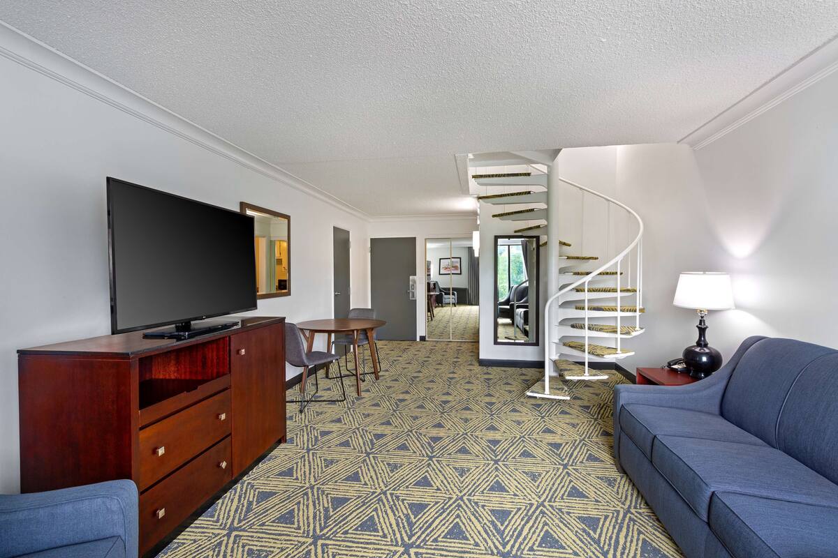 A modern living room with a flat-screen TV, a wooden cabinet, a blue sofa, a patterned carpet, and a spiral staircase leading to another room.