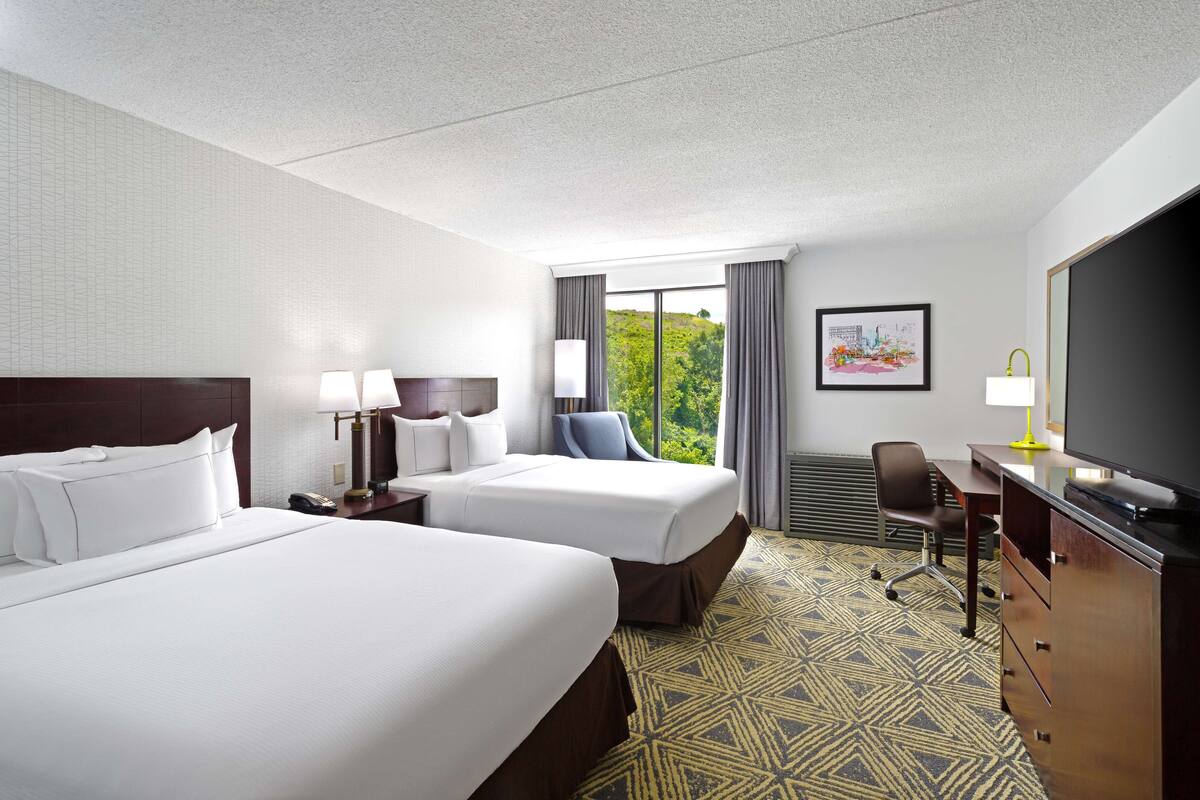 A hotel room with two beds, a desk with a TV, a chair, and a window with a view of greenery.