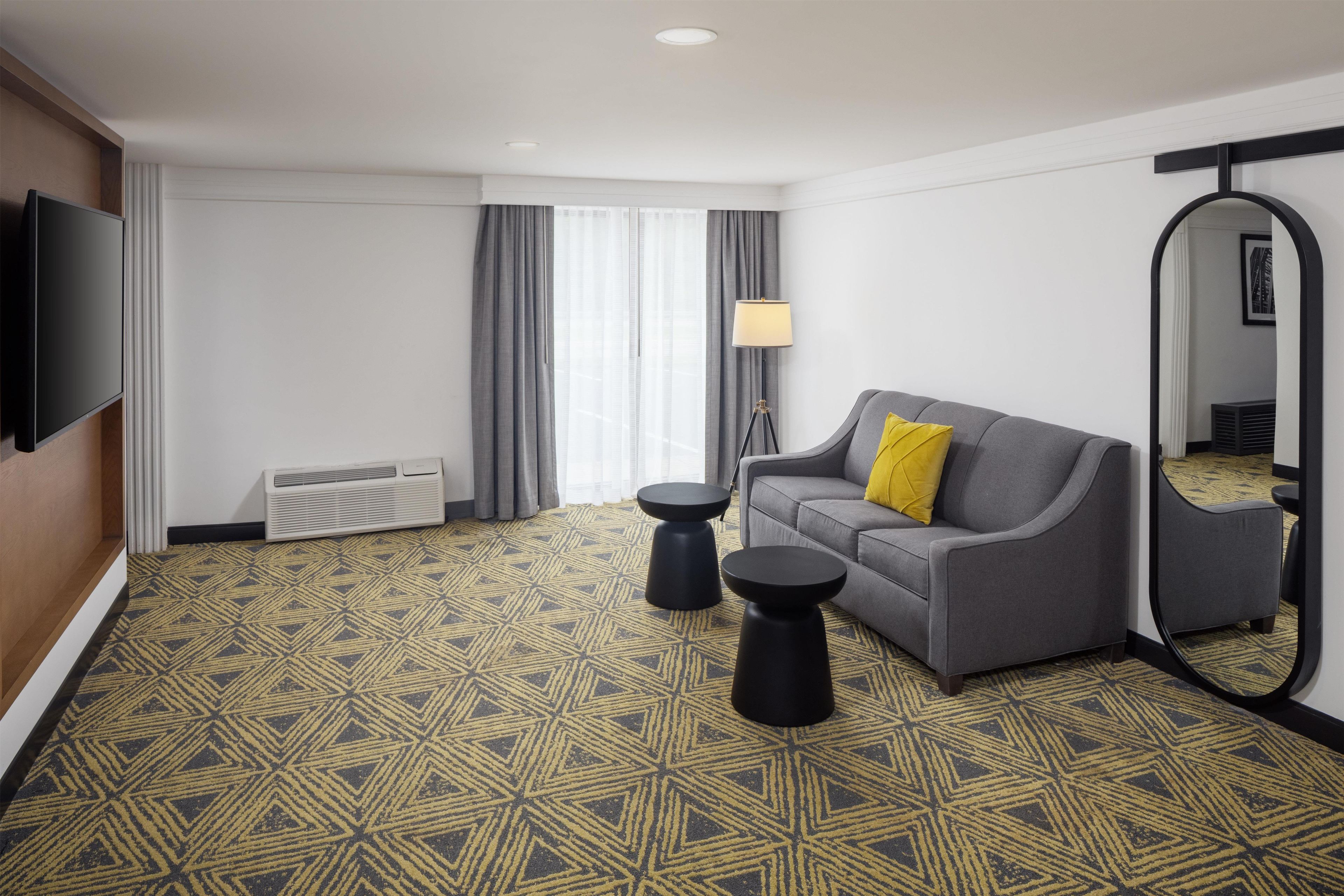 A hotel room with a grey sofa, a yellow pillow, a round black side table, a floor lamp, a large mirror, and a patterned carpet.