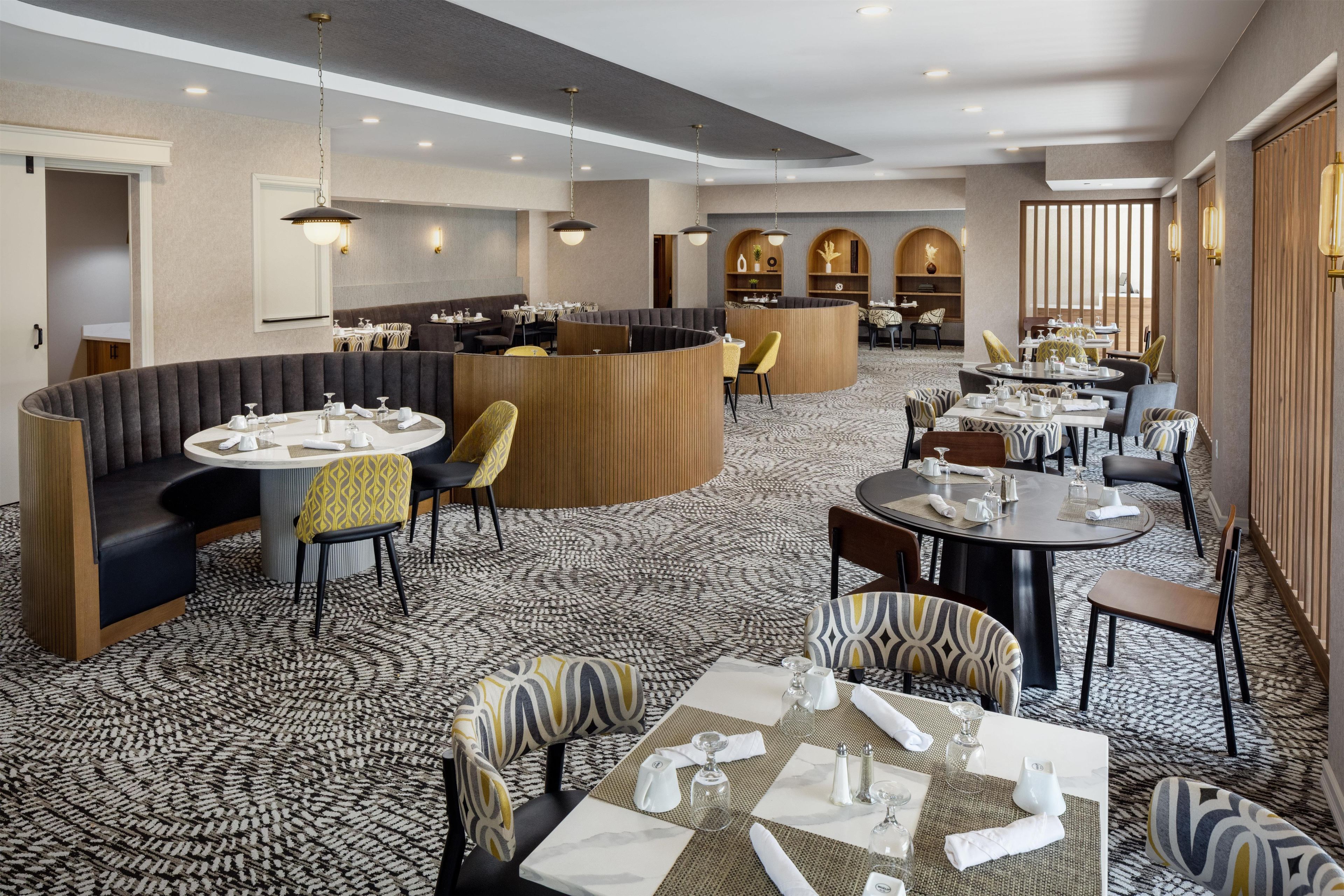 A hotel dining area with round tables, cushioned seating, and a patterned carpet.