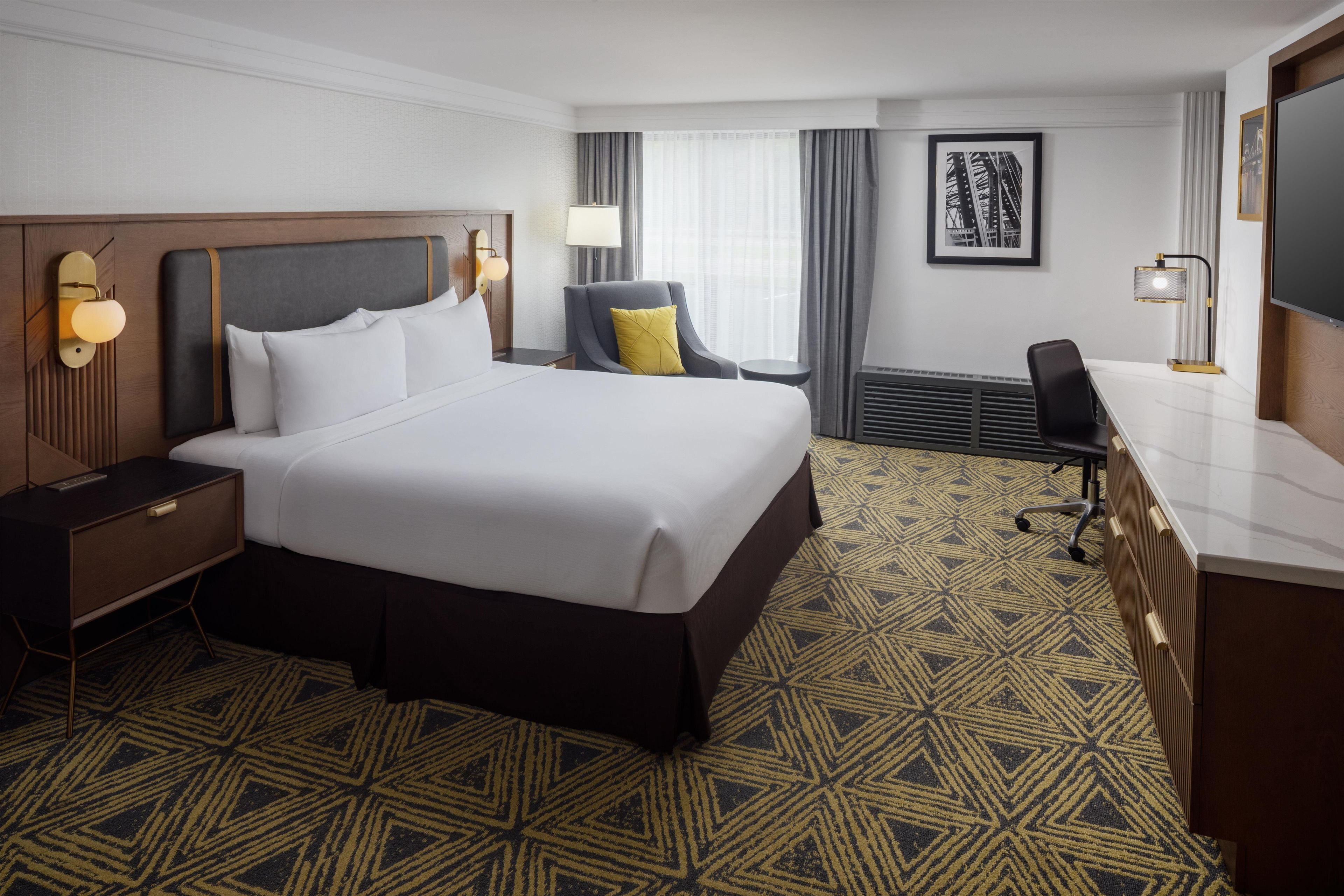 A hotel room with a large bed, a desk, a chair, a nightstand, a lamp, a picture on the wall, and a patterned carpet.