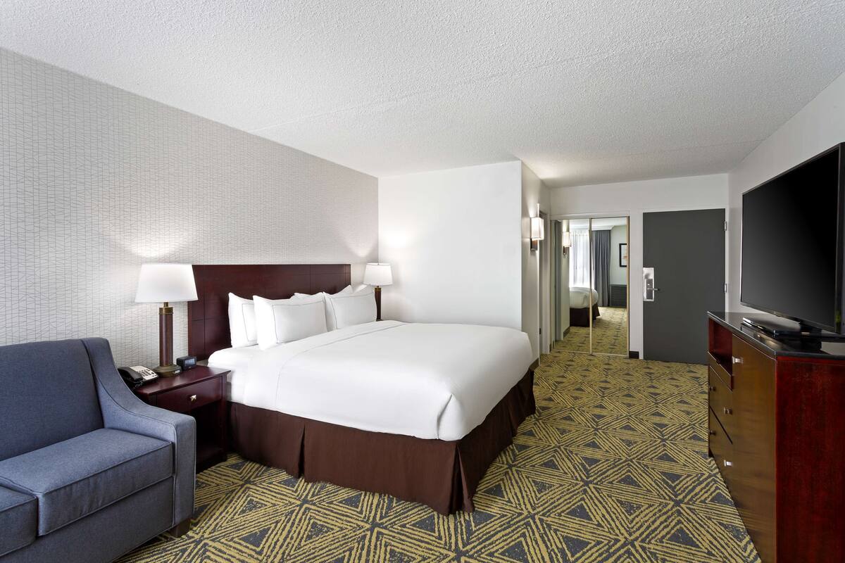 A hotel room with a large bed, a desk with a lamp, a television, and a patterned carpet.