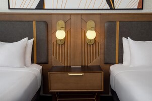 Habitación de hotel con dos camas, una mesita de noche con cajón e iluminación en la pared.