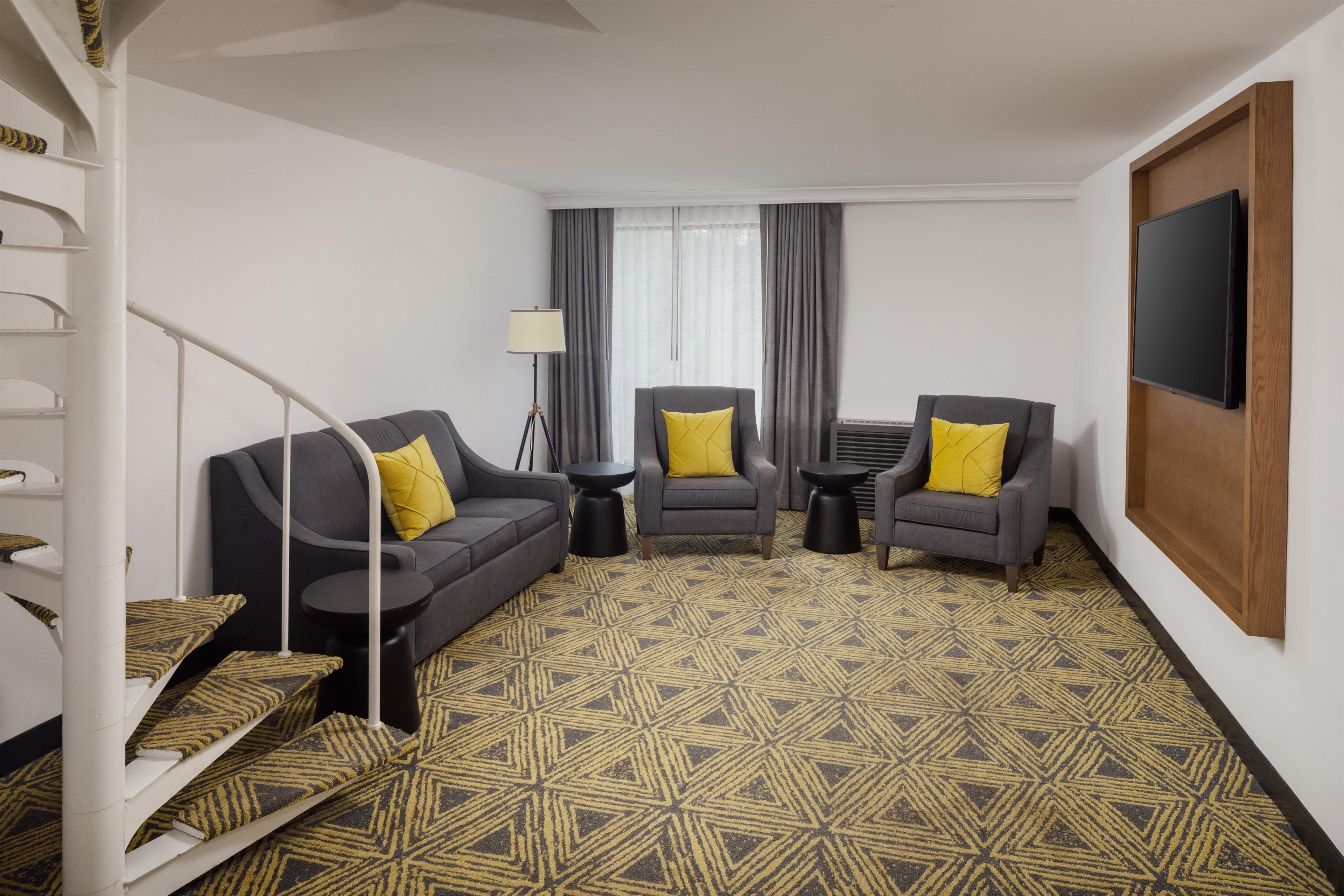 A hotel room with a grey sofa, two armchairs, a small table, a floor lamp, a TV, and a patterned carpet.