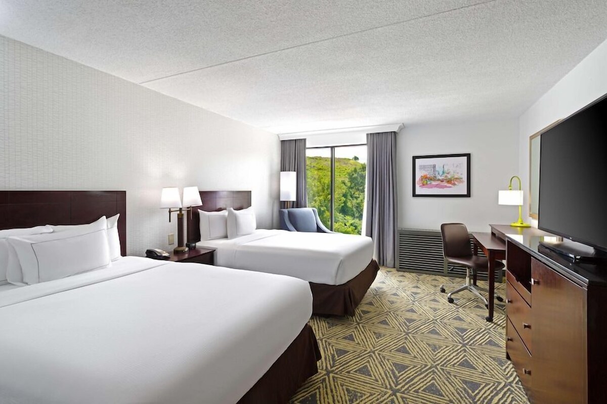 A hotel room with two beds, a desk, a chair, a television, and a window with a view of greenery.