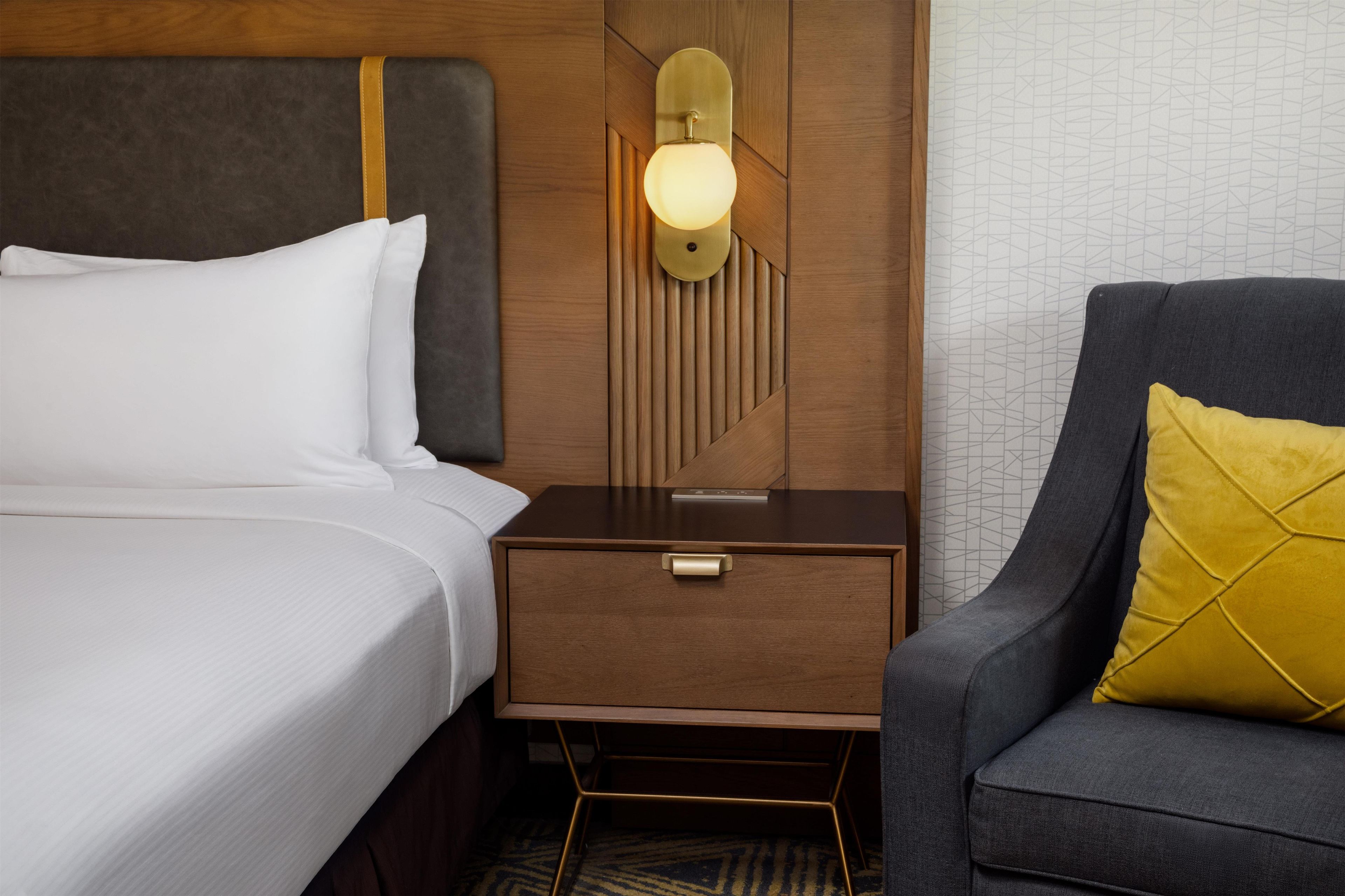 A hotel room with a bed, a nightstand with a lamp, and a seating area with a chair and a pillow.