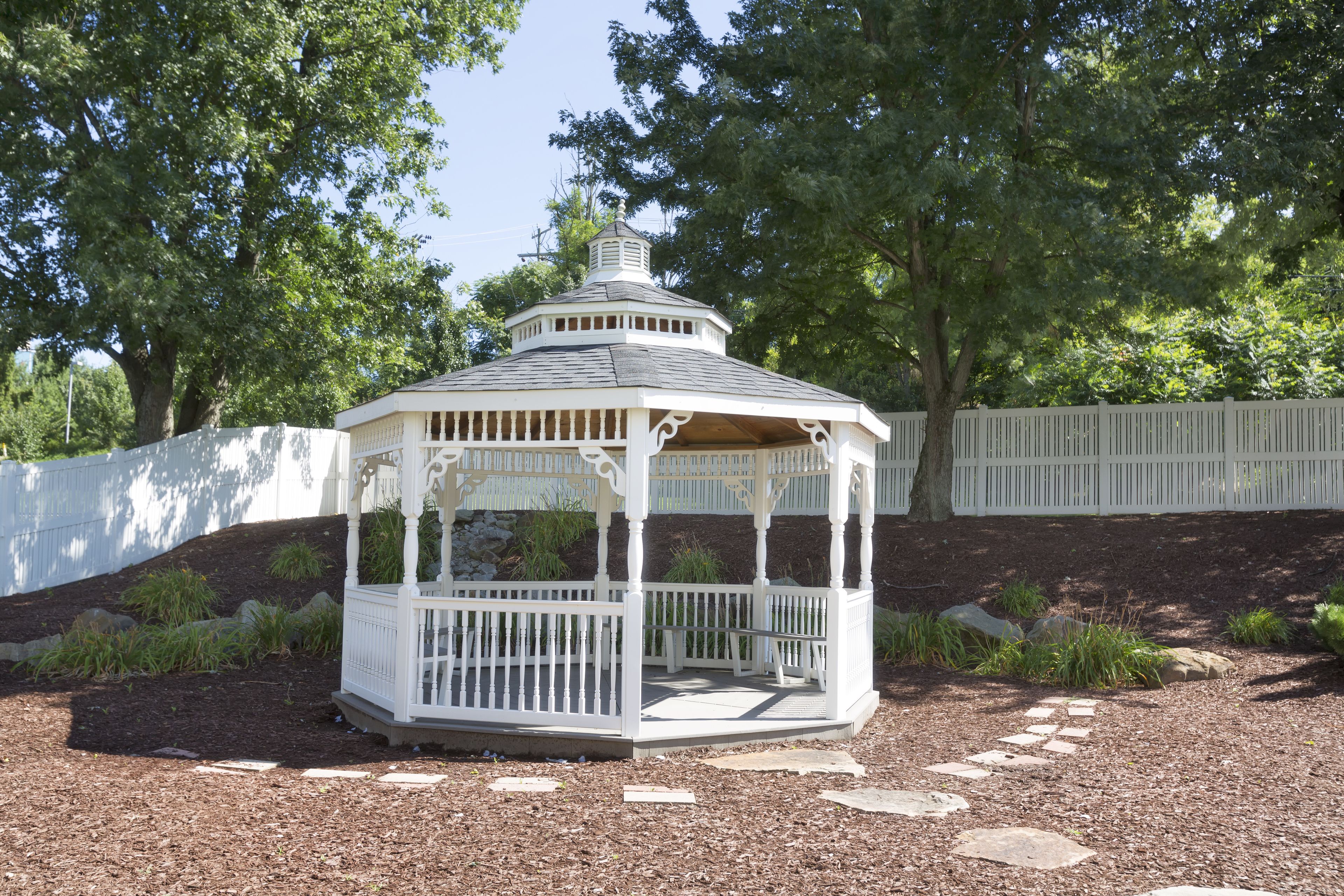 A gazebo with a white wooden structure and a roof, surrounded by a gravel path and greenery.