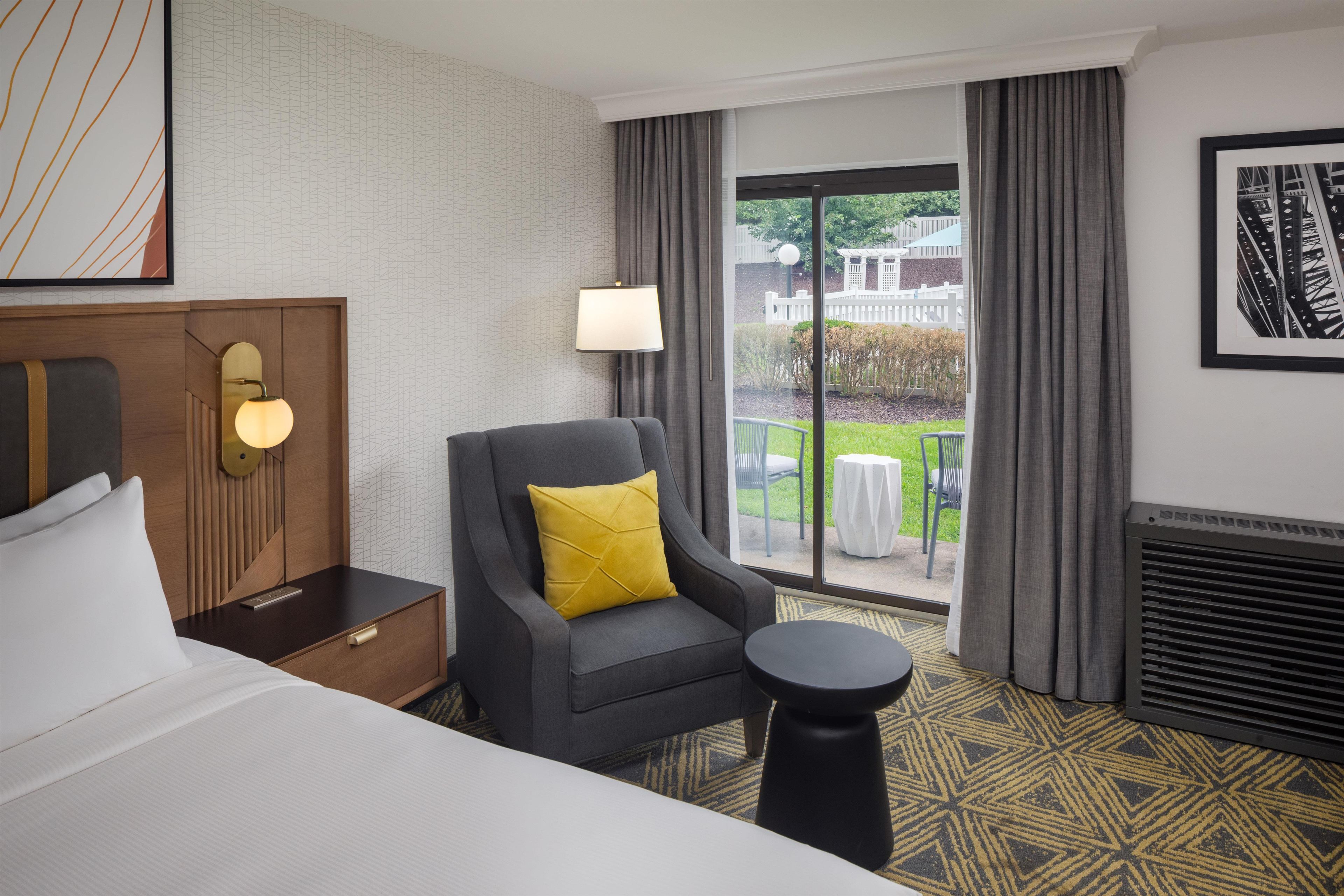 A hotel room with a bed, a chair with a yellow pillow, a nightstand, a lamp, and a view of the outdoors through a large window.