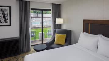 A hotel room with a bed, a chair with a yellow pillow, a small table, and a view of a pool and white fence.