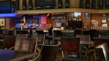Un espace bar avec différents types de sièges, un téléviseur fixé au mur et diverses bouteilles d’alcool exposées sur des étagères.