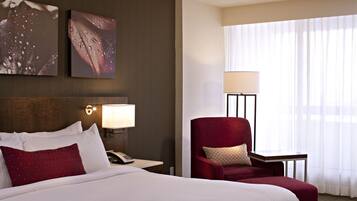 A hotel room with a large bed, a desk with a lamp, a red armchair, and two wall-mounted artworks.