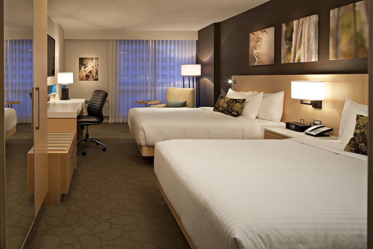 Quarto de hotel com duas camas, uma escrivaninha, uma cadeira e uma mesinha.
