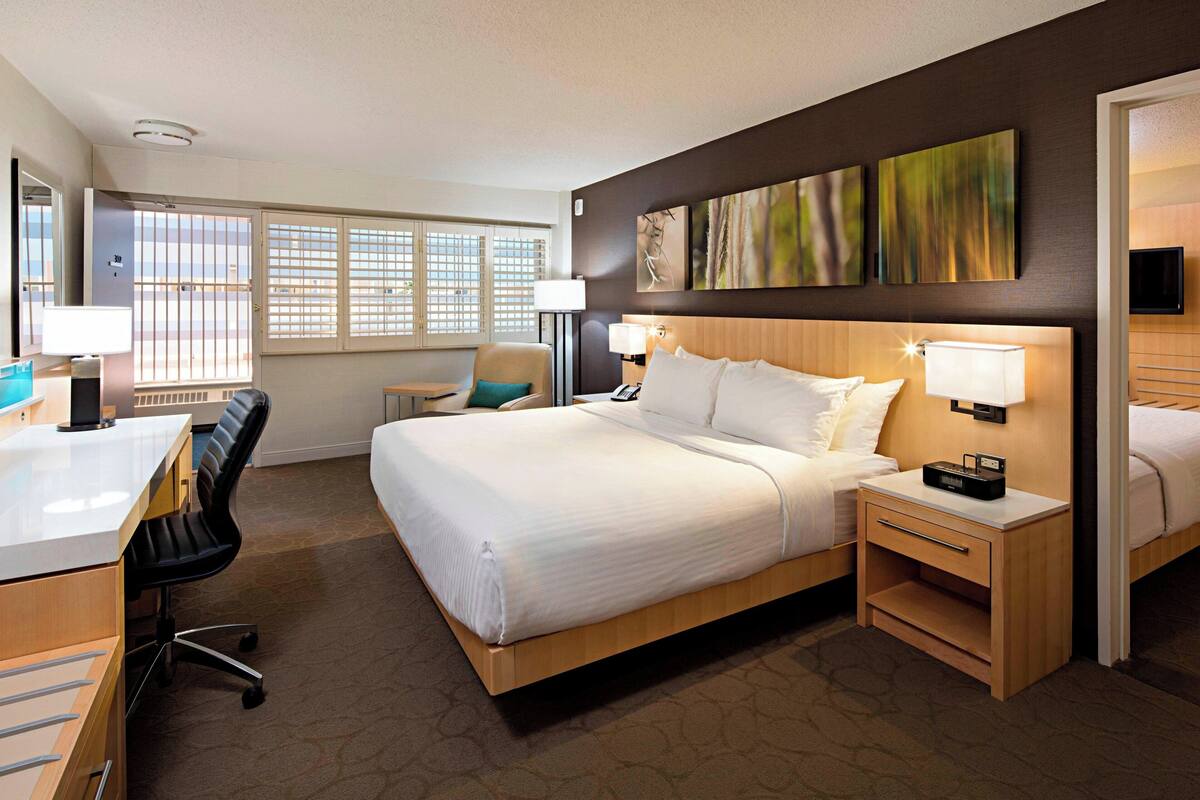 Quarto de hotel com uma cama grande, uma escrivaninha, uma cadeira, uma mesinha de cabeceira e uma televisão.