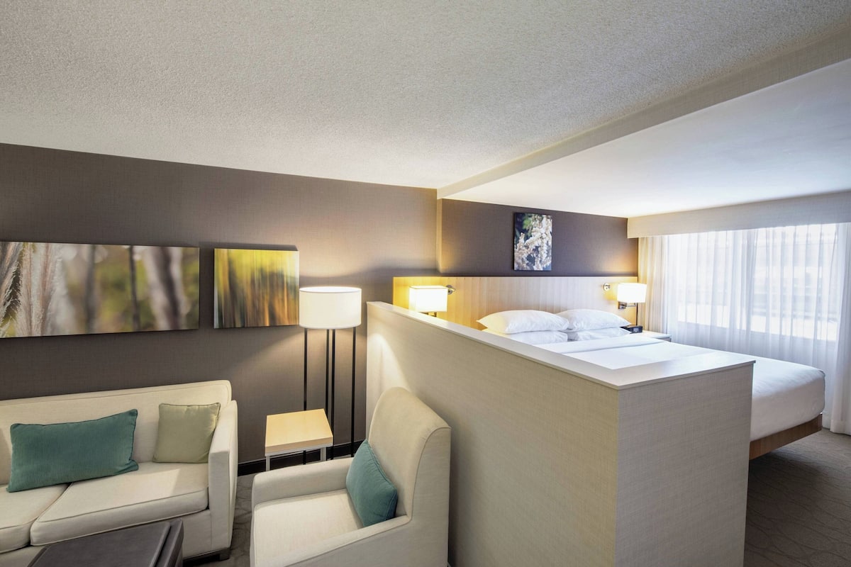 Quarto de hotel com uma cama, um sofá, uma cadeira, uma escrivaninha e duas luminárias fixadas na parede.